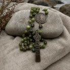 An olive green beaded rosary with antique bronze-toned metal beads, a round medallion, and a detailed cross pendant, arranged on draped beige fabric with a smooth gray stone behind it.