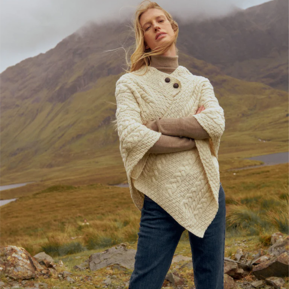 A woman stands outdoors in a grassy mountain landscape wearing a cream cable-knit poncho-style sweater over a taupe turtleneck, with dark blue jeans.