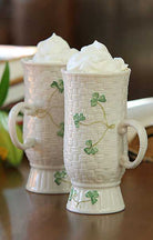 Two ceramic mugs with shamrock designs filled with whipped cream on a wooden surface.