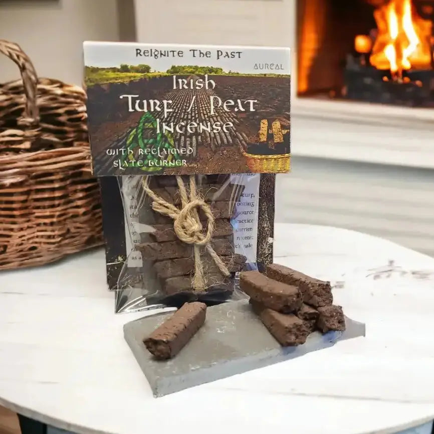 A package of Irish Tarf / Peat Incense by Auralc, displayed on a white table with a blurred fireplace in the background. The clear bag contains dark brown peat briquettes tied with a jute twine, with a few briquettes placed on a gray stone slab in front of the package.