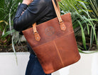 Brown leather tote bag with Celtic knot design held by a person outdoors.