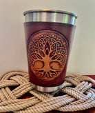 Brown leather cup with tree of life design on a metal sleeve, placed on woven mat.