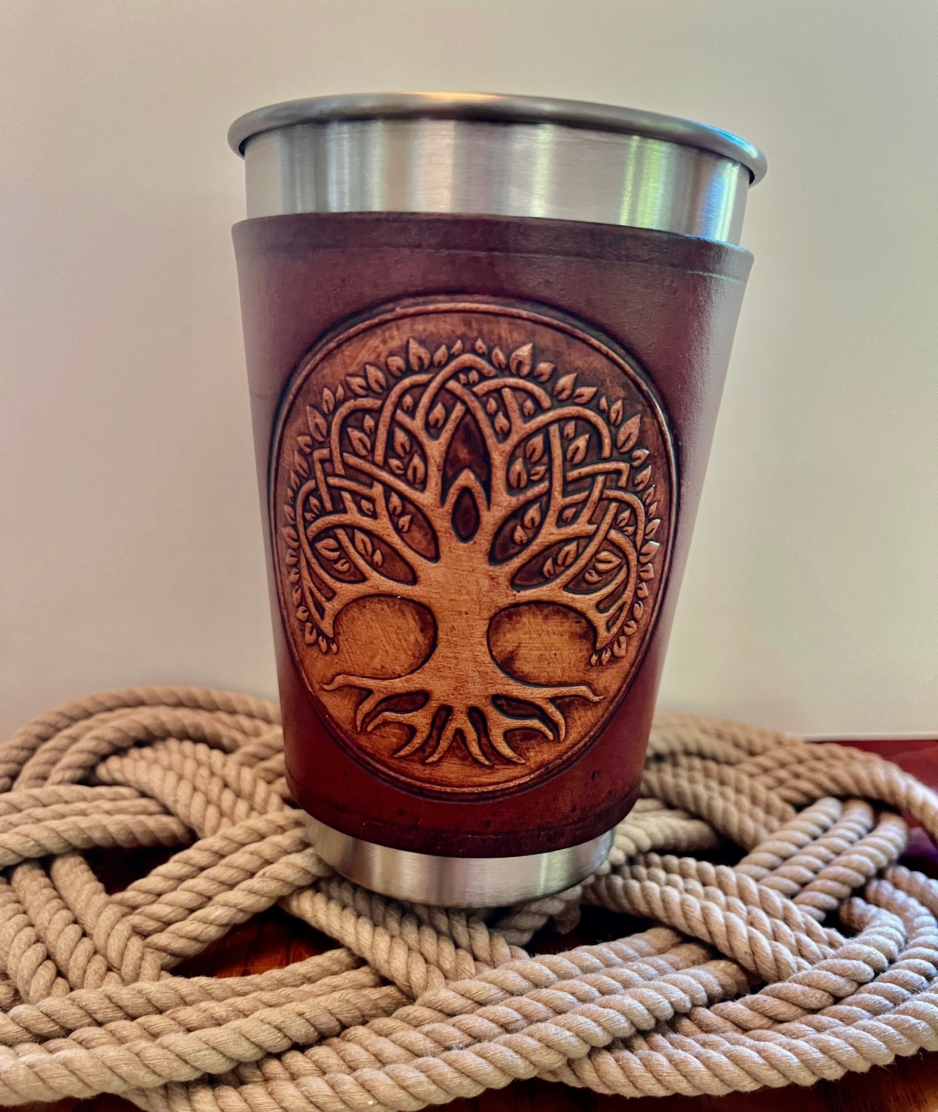 Brown leather cup with tree of life design on a metal sleeve, placed on woven mat.