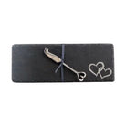 A silver metal cheese knife with a heart-shaped handle, presented on a dark gray slate card tied with a navy ribbon, featuring an engraved double heart design.