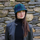 Woman wearing a plaid hat and black jacket against a stone wall.
