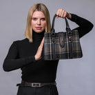 A woman in a black turtleneck sweater holds up a structured plaid handbag with black leather handles and trim. The handbag features a brown, black, and beige tartan pattern with gold-tone hardware.