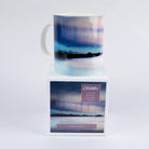 A ceramic mug with a landscape print of Scottish aurora (blue, purple, and black mountains over water) sits on top of its white retail packaging box. The box features the Cath Waters  logo and product details.