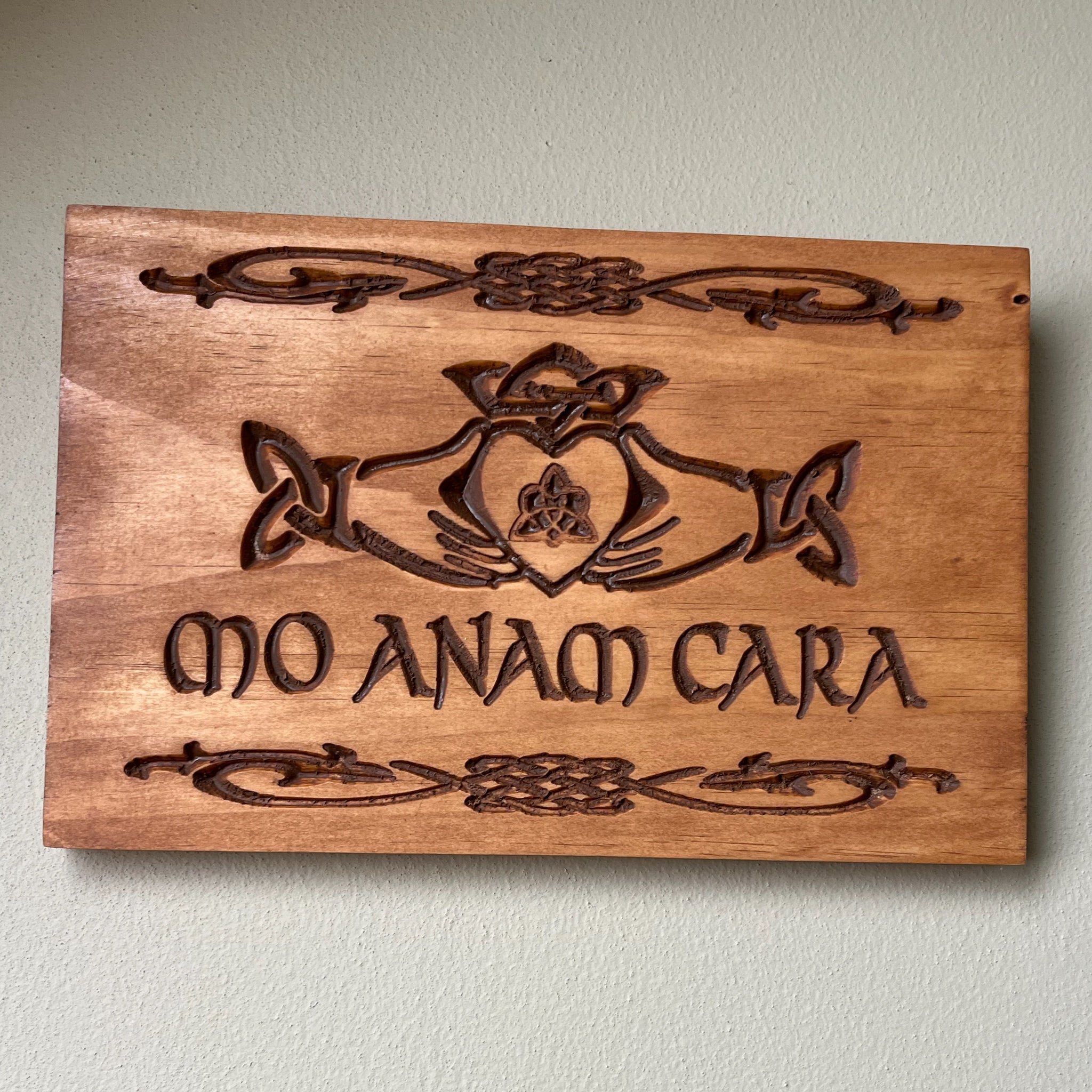 A rectangular wooden plaque with hand-carved Celtic knotwork and the Latin phrase 'MO ANAM CARA' in the center, mounted on a light-colored textured wall.