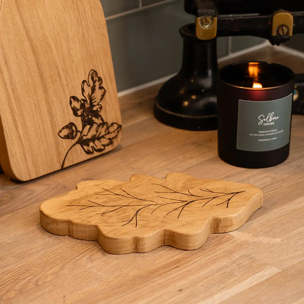 A leaf-shaped wooden coaster with engraved leaf veins sits on a wooden tabletop next to a lit candle and another wooden board with a burned floral design.