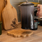A dark gray to black gradient enamel coffee pot with a silver lid and handle, held by a hand on a wooden kitchen countertop. In the foreground, a leaf-shaped wooden trivet sits on the counter, with a wooden cutting board and other kitchen items in the background.