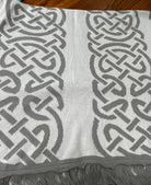A close-up of a woven scarf or throw blanket with a gray and white Celtic knot pattern and fringed edges, laid flat on a wooden surface.