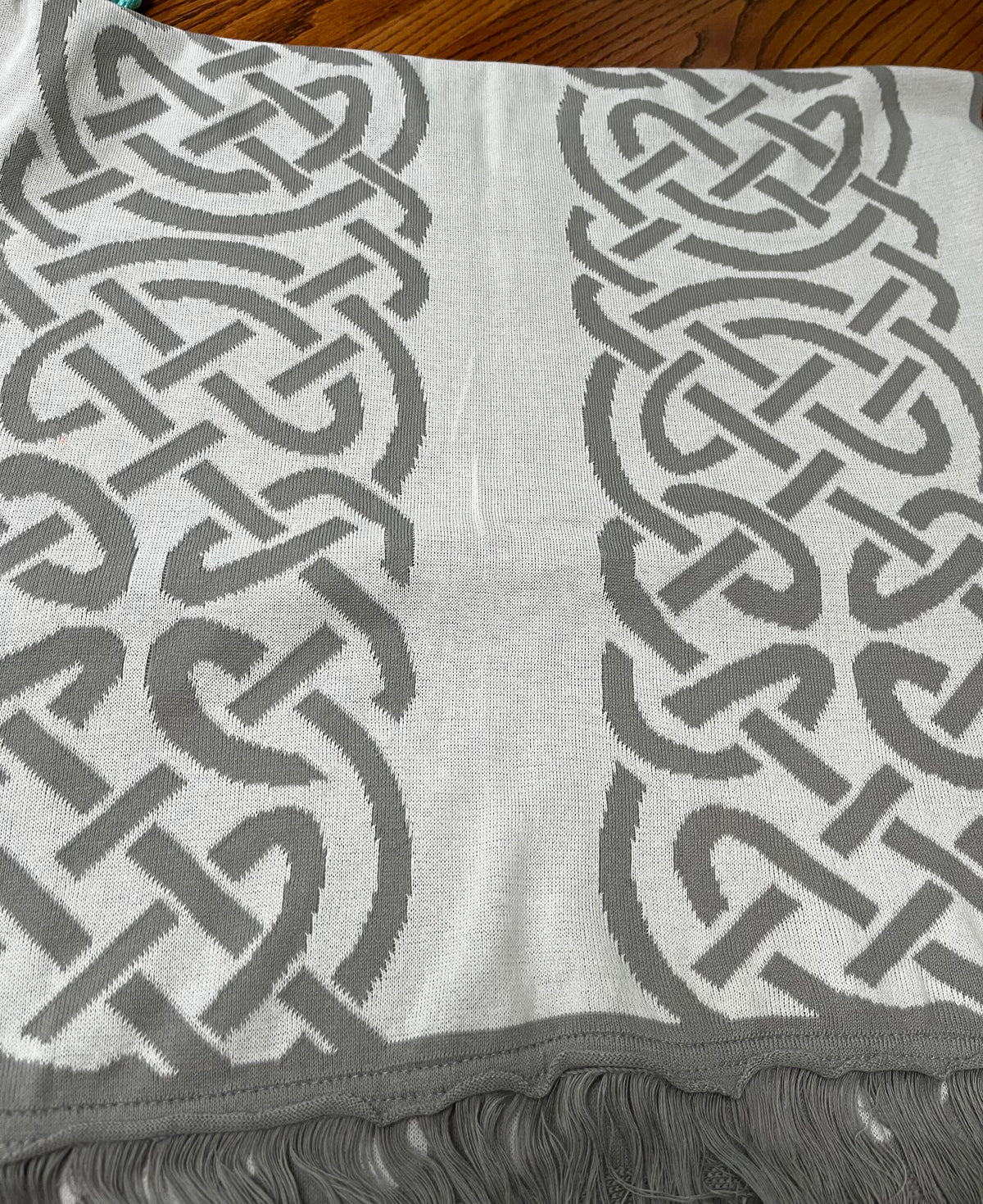 A close-up of a woven scarf or throw blanket with a gray and white Celtic knot pattern and fringed edges, laid flat on a wooden surface.