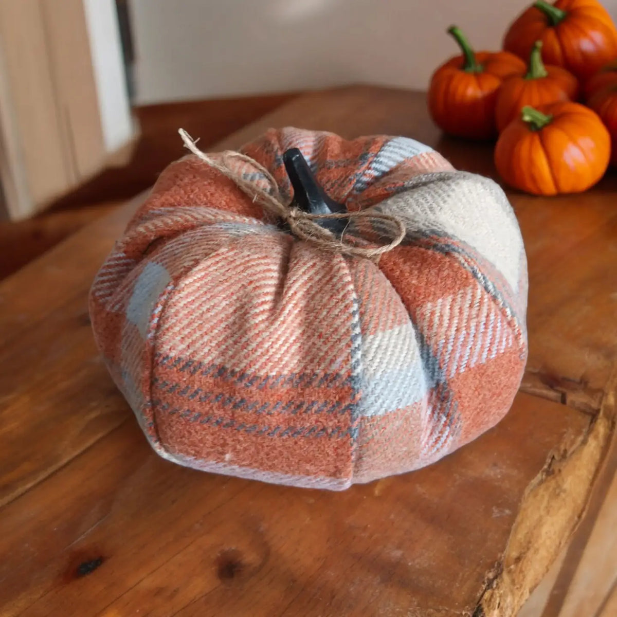A handmade fabric pumpkin decoration made from a plaid wool-like material in muted orange, beige, and gray tones, with a small black stem and twine bow, displayed on a wooden table with small orange pumpkins in the background.