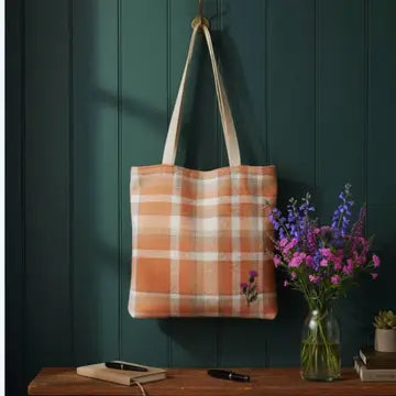 A canvas tote bag with a peach and white plaid pattern hangs on a dark teal wooden wall. The bag has beige shoulder straps and a small embroidered purple flower detail near the bottom right.
