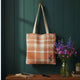 A canvas tote bag with a peach and white plaid pattern hangs on a dark teal wooden wall. The bag has beige shoulder straps and a small embroidered purple flower detail near the bottom right.