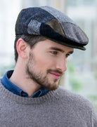 A man wearing a textured patchwork flat cap in gray, black, and brown herringbone patterns, styled with a blue collared shirt and a gray knit sweater.