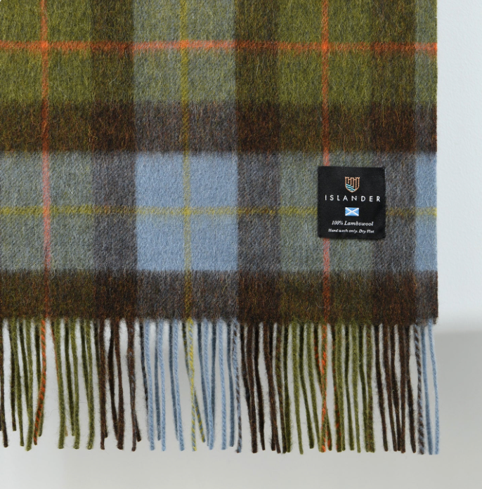A close-up of a plaid blanket with fringe, featuring olive green, brown, gray, and light blue checks with thin orange and yellow lines. A black woven label reading ISLANDER is sewn onto the right side.