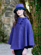 A woman wearing a textured purple cape with three black buttons and a matching purple hat with a black brim, standing outdoors against a stone wall and green foliage.