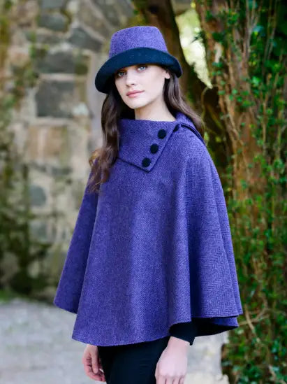 A woman wearing a textured purple cape with three black buttons and a matching purple hat with a black brim, standing outdoors against a stone wall and green foliage.