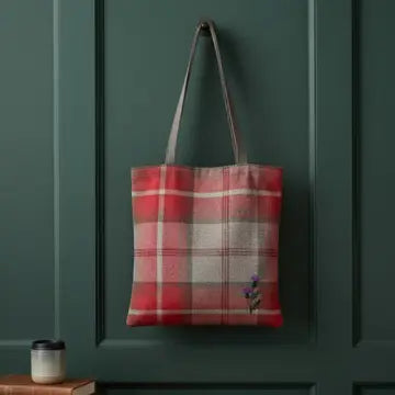 A red and beige plaid canvas tote bag with brown shoulder straps hangs on a dark green paneled door. A small purple flower is embroidered near the bottom right corner of the bag.