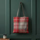 A red and beige plaid canvas tote bag with brown shoulder straps hangs on a dark green paneled door. A small purple flower is embroidered near the bottom right corner of the bag.