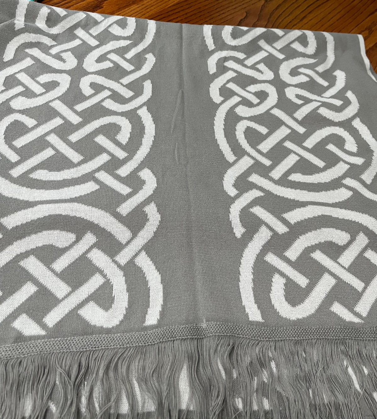 A gray knit scarf with a white Celtic knot pattern and fringed edges, laid flat on a wooden surface.