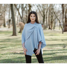 A woman wearing a light blue wrap poncho over a pink long-sleeve top and black pants, standing in a park with blurred trees in the background.