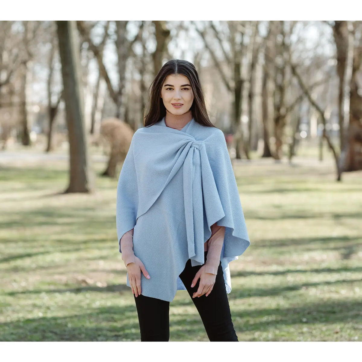 A woman wearing a light blue wrap poncho over a pink long-sleeve top and black pants, standing in a park with blurred trees in the background.