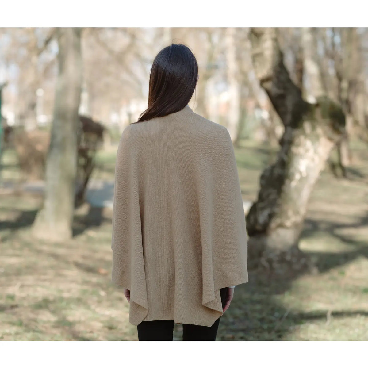 A woman viewed from behind wearing a light beige open-front poncho or cape in an outdoor park setting with blurred trees.
