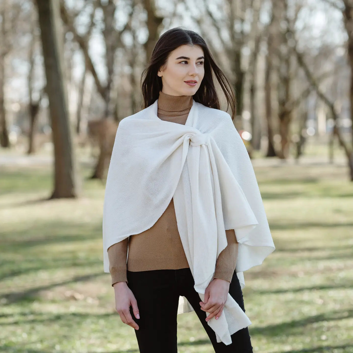 A woman wearing a cream-colored knit wrap or poncho tied around her shoulders, styled over a brown turtleneck and black pants, standing in a park with blurred trees in the background.