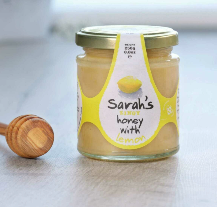 Jar of 'Sarah's Honey with Lemon' on a light surface with a wooden honey dipper.