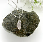 Silver leaf-shaped pendant on a chain, resting on a textured stone surface with white flowers in the background.