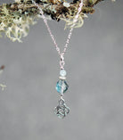 Necklace with a pendant featuring a Celtic knot design and a blue gemstone, hanging from a branch with lichen.