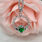 A silver Claddagh heart-shaped pendant necklace with a green heart gemstone, displayed against soft pink rose petals.