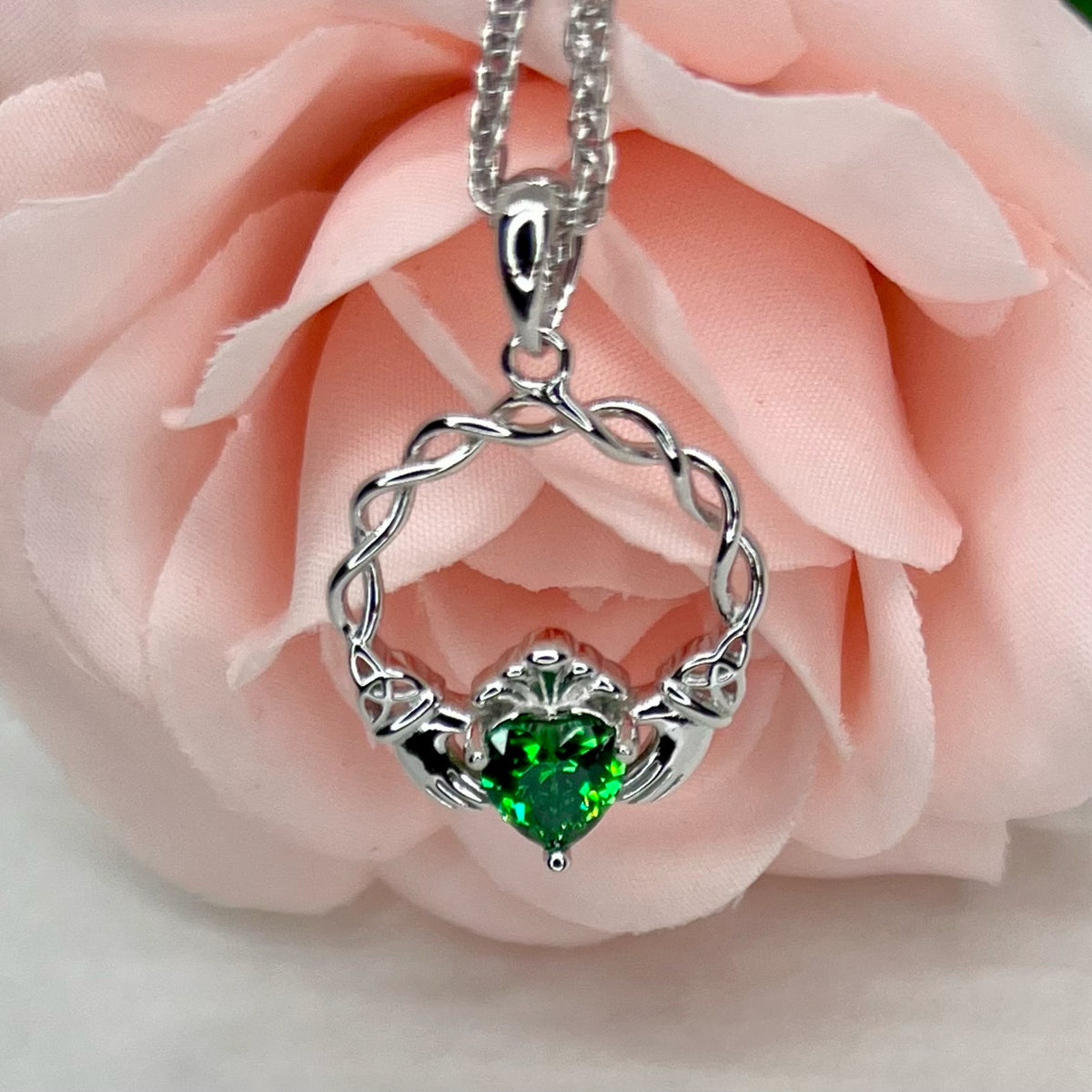 A silver Claddagh heart-shaped pendant necklace with a green heart gemstone, displayed against soft pink rose petals.