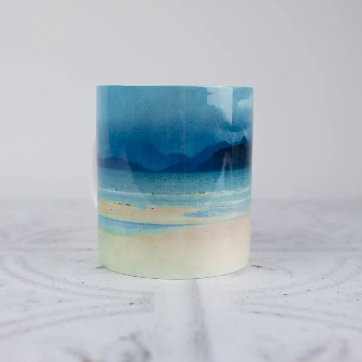 A ceramic mug with watercolor beach landscape print, featuring shades of blue, beige, and white, set against a light gray background.