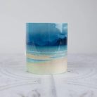 A ceramic mug with watercolor beach landscape print, featuring shades of blue, beige, and white, set against a light gray background.