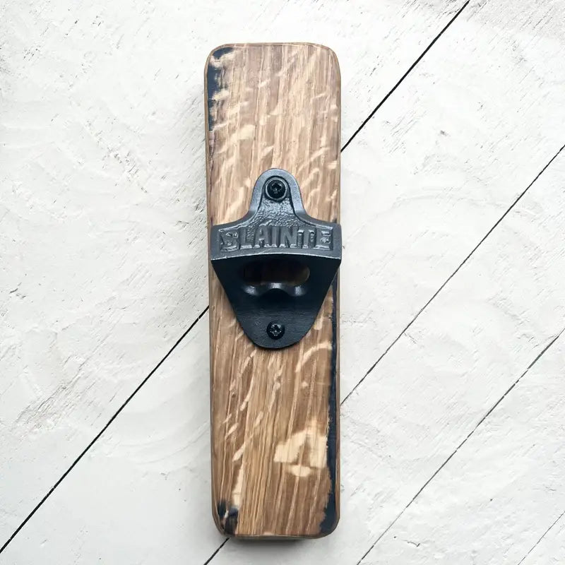 A rustic wall-mounted bottle opener featuring a rectangular stained wood block with a black metal blade. The blade is embossed with the brand name 'BLAINDE' and is mounted on a white-painted wooden plank background.
