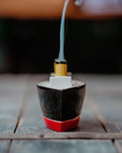 A small decorative object shaped like a ship's bow, with a black upper section, a red base, a white rectangular detail, and a yellow top, shown on a wooden surface with a blurred background and a thin blue-gray smoke-like streak rising from it.