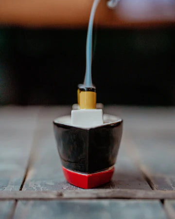 A small decorative object shaped like a ship's bow, with a black upper section, a red base, a white rectangular detail, and a yellow top, shown on a wooden surface with a blurred background and a thin blue-gray smoke-like streak rising from it.