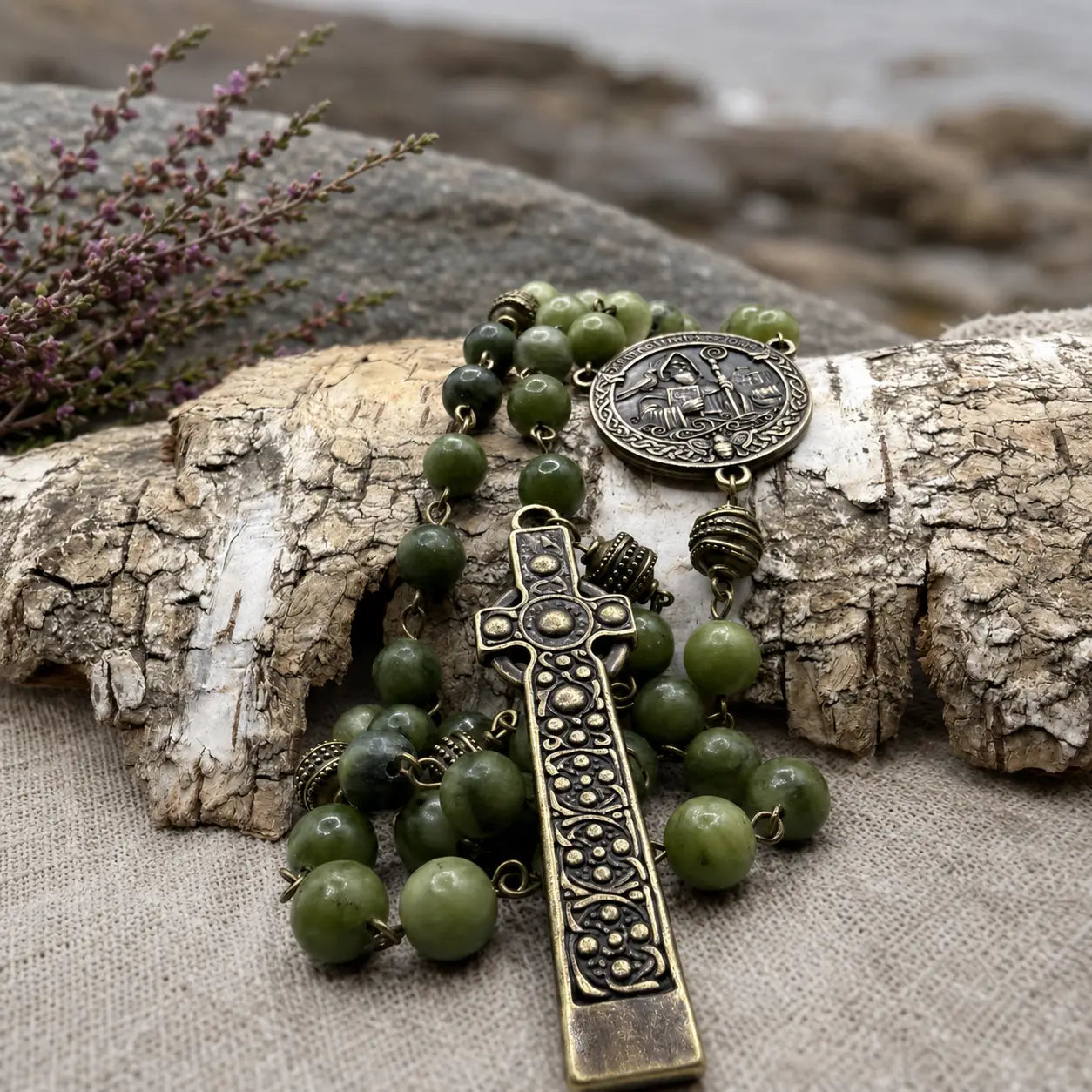 An olive green beaded rosary with an ornate antique bronze cross and medallion, styled on a piece of weathered birch wood with a linen surface and blurred natural background.