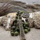 An olive green beaded rosary with an ornate antique bronze cross and medallion, styled on a piece of weathered birch wood with a linen surface and blurred natural background.