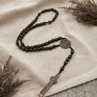 A dark brown beaded rosary with a silver-toned metal centerpiece medallion and a matching cross pendant, arranged on draped cream linen fabric with dried floral sprigs in the corners.