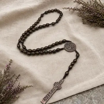 A dark brown beaded rosary with a silver-toned metal centerpiece medallion and a matching cross pendant, arranged on draped cream linen fabric with dried floral sprigs in the corners.