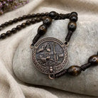 A close-up of a beaded necklace with dark brown wooden beads and a round bronze-toned pendant featuring an engraved figure, displayed on a smooth gray stone over beige fabric.