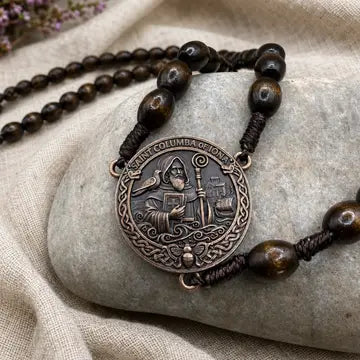 A close-up of a beaded necklace with dark brown wooden beads and a round bronze-toned pendant featuring an engraved figure, displayed on a smooth gray stone over beige fabric.