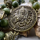 A close-up of a Saint Patrick beaded necklace featuring an ornate bronze-toned medallion pendant with an embossed figure and Celtic knotwork, surrounded by round green beads and decorative metal spacers, displayed on a textured wood surface.