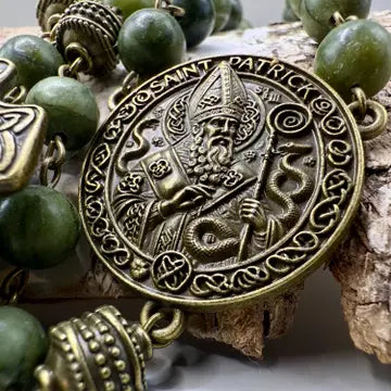 A close-up of a Saint Patrick beaded necklace featuring an ornate bronze-toned medallion pendant with an embossed figure and Celtic knotwork, surrounded by round green beads and decorative metal spacers, displayed on a textured wood surface.