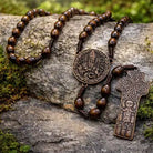 A wooden and metal religious rosary with a round medallion and a cross pendant, displayed on a mossy rock outdoors.