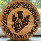 A round wooden plaque or coaster featuring a Celtic knot border and a central engraved thistle design, photographed in front of a window.